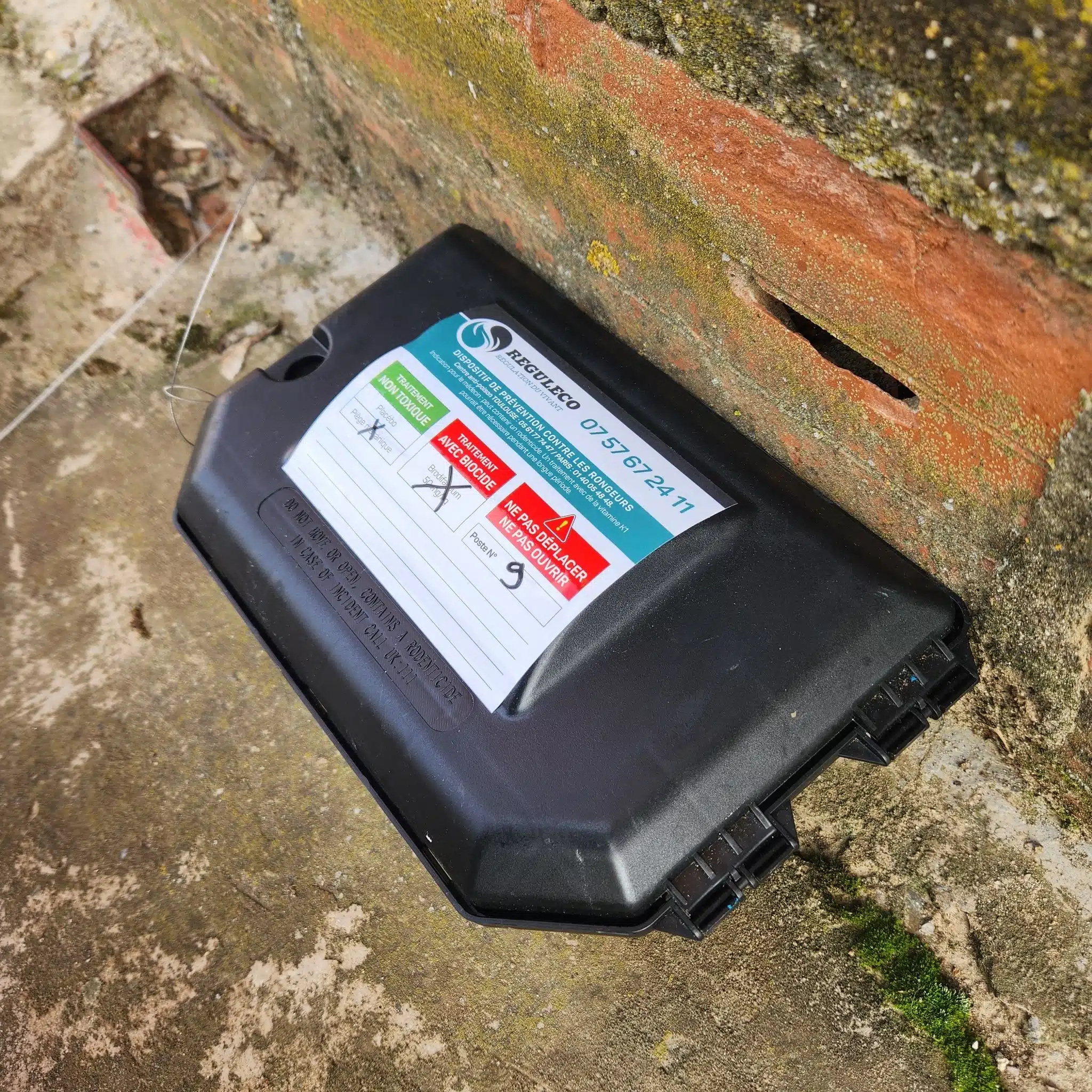 Poste de dératisation noir Réguleco (Poste N° 9) sur sol extérieur. Contient du biocide Brodifacoum. Ne pas déplacer ni ouvrir.
